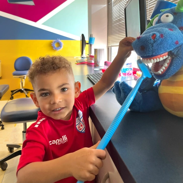 Young boy in a red shirt smiling and holding a large toothbrush at the dental office